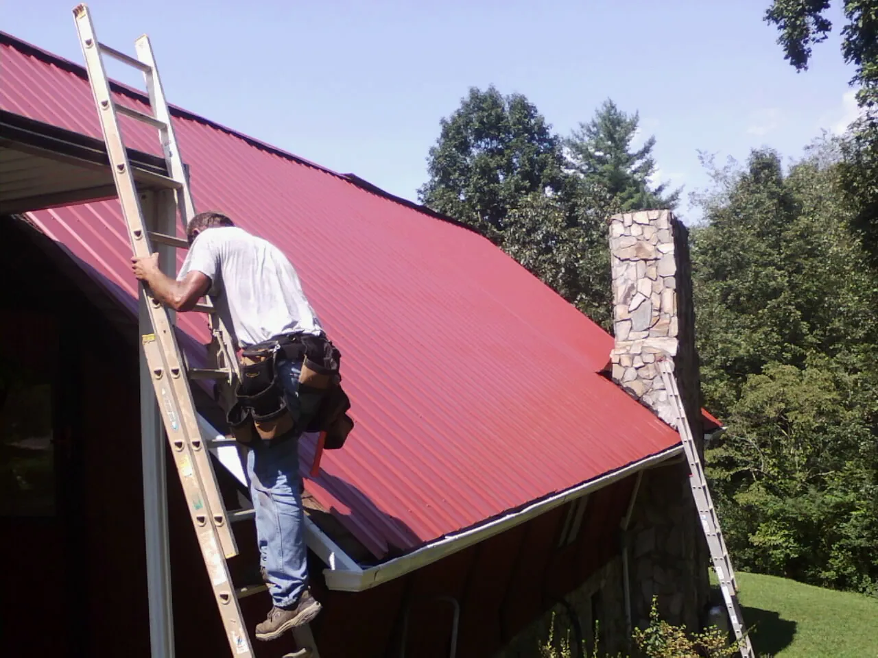 Roofer on a ladder beside a red metal roof and stone chimney — Soffit & Fascia Repair by Roofing Brothers in Lake Zurich